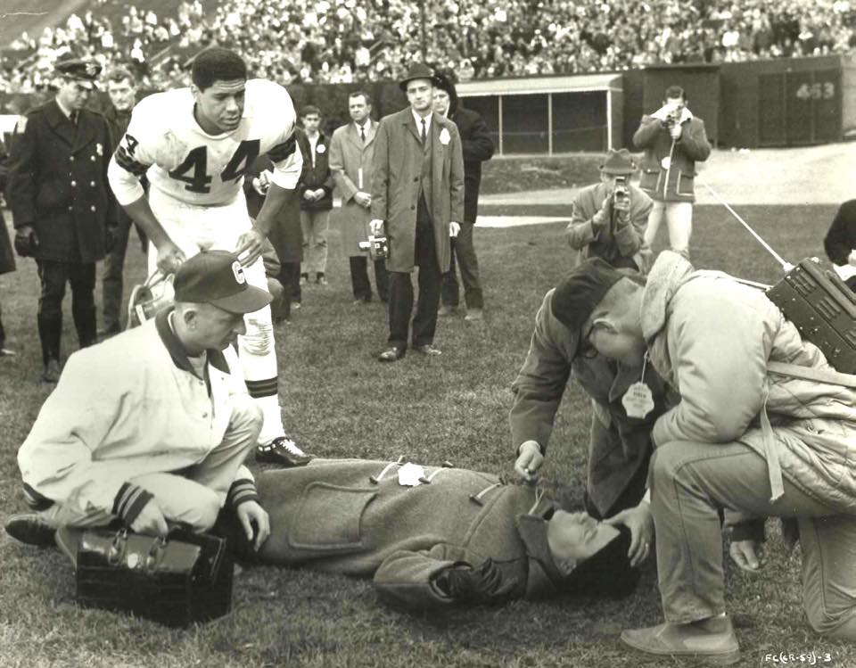 Marty kneeling on a Cleveland Browns field with his medical kit, attending to a player, with #44 standing nearby