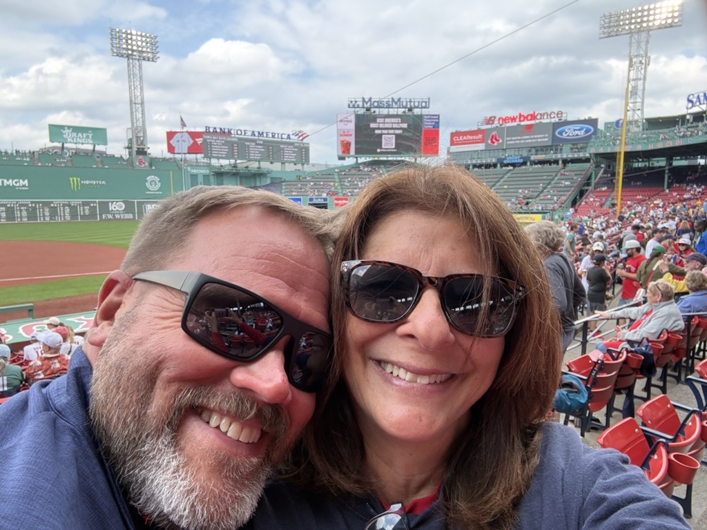 Mum and Eric at Fenway Park, smiling in sunglasses with the Green Monster behind